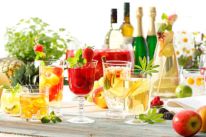 verschiedene Frucht-Bowlen in unterschiedlichen Gläsern auf einem Tisch mit Wein- und Sektflaschen und Früchten als Dekoration