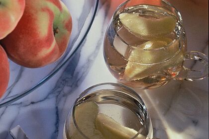 Pfirsichbowle in zwei Gläsern auf einem Marmortisch, daneben eine Obstschale mit Pfirsichen und ein Bowlegefäß.